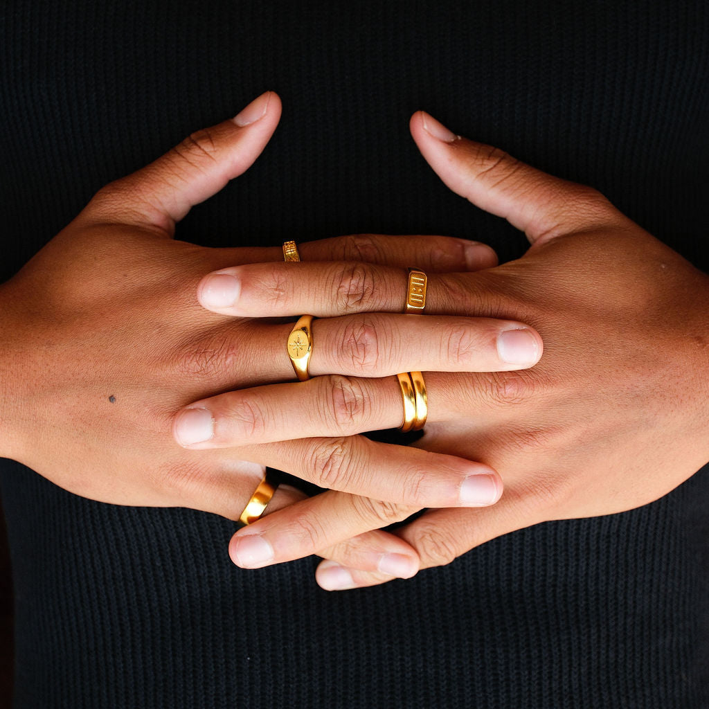 A man's hands clasped, showing off a collection of gold rings against a black knit jumper. The Billie Jo Luminous Ring is visible on the middle finger.