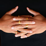 A man's hands clasped, showing off a collection of gold rings against a black knit jumper. The Billie Jo Luminous Ring is visible on the middle finger.