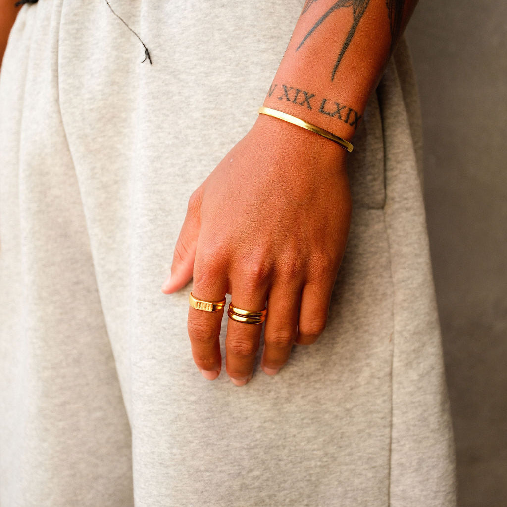 A man's hand, resting against light-coloured trousers, wearing a stack of gold rings from Billie Jo. The image highlights the versatility of the Stacker Ring for everyday wear.