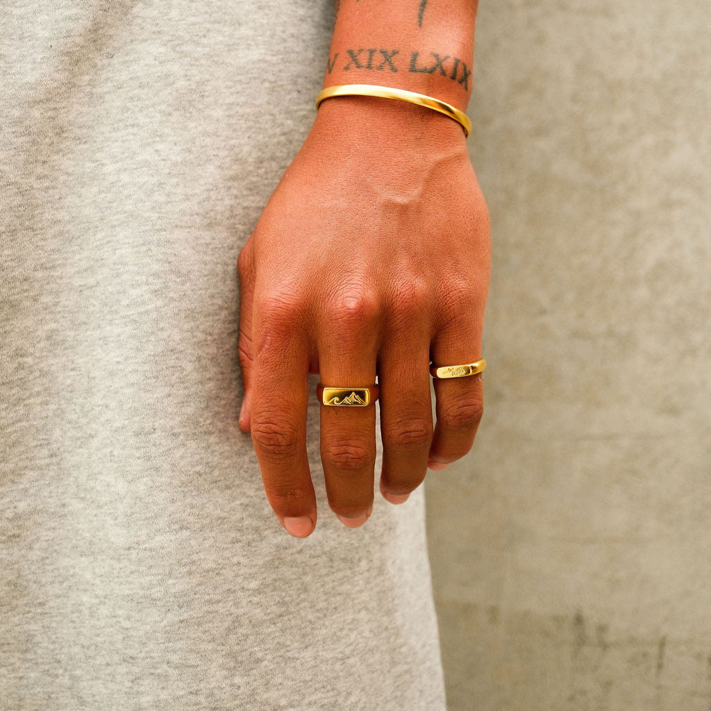 A man with a tattooed wrist wears the Billie Jo Rosa II Ring and other gold jewellery, showcasing the brand's modern and masculine aesthetic.