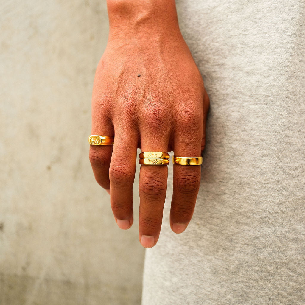 A close-up of a man's hand wearing the Billie Jo Koi I Ring and other gold rings against a grey marle fabric. A modern, casual look for men's jewellery.