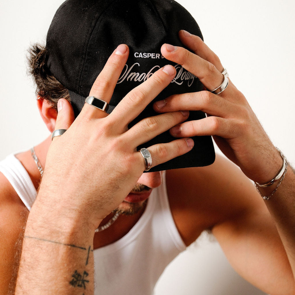 A man in a white tank top and black cap, with his hands covering his face, wearing the Revival Ring and other Billie Jo men's jewellery.