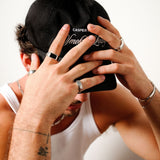 A man in a white tank top and black cap, with his hands covering his face, wearing the Revival Ring and other Billie Jo men's jewellery.
