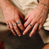 A man's hands with several silver rings, including the Serenity Ring, resting on his lap. He is wearing beige trousers.
