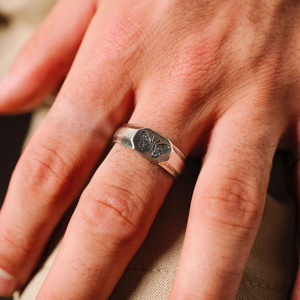 A close-up of the Revival Ring on a man's finger, showing the detail of the butterfly engraving and the textured band. Part of the Billie Jo collection.