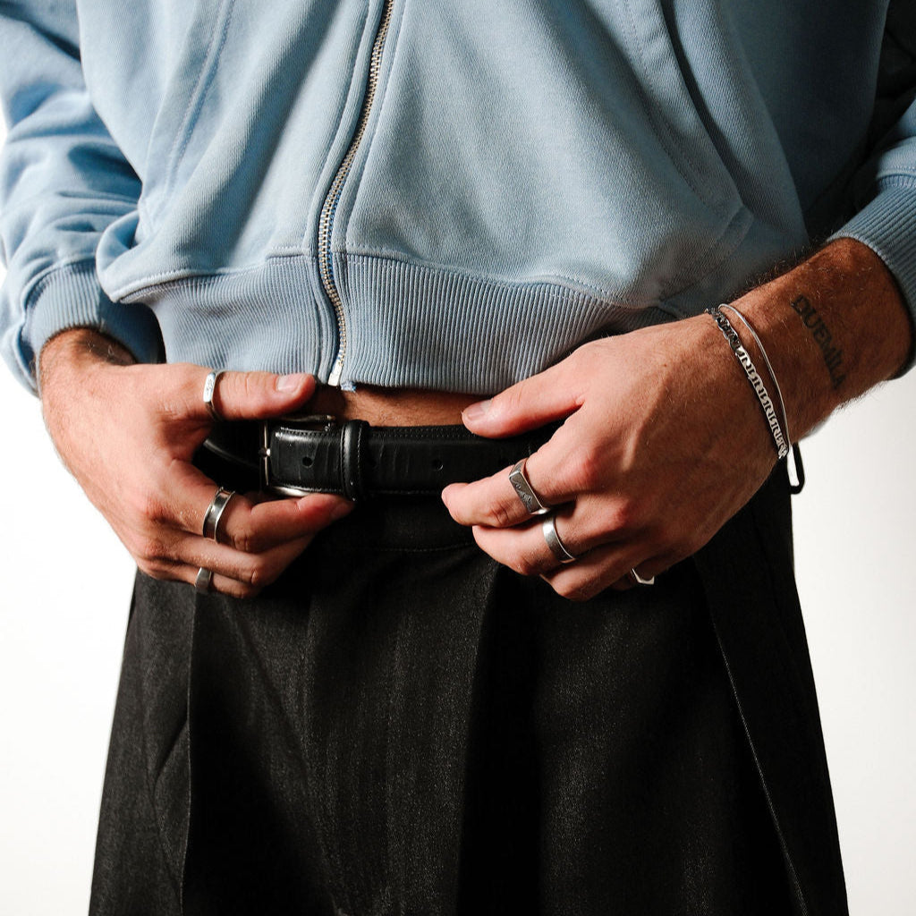 A man wearing a blue zip-up hoodie and black trousers adjusts his belt, his hands feature the Serenity Ring and other silver rings by Billie Jo.