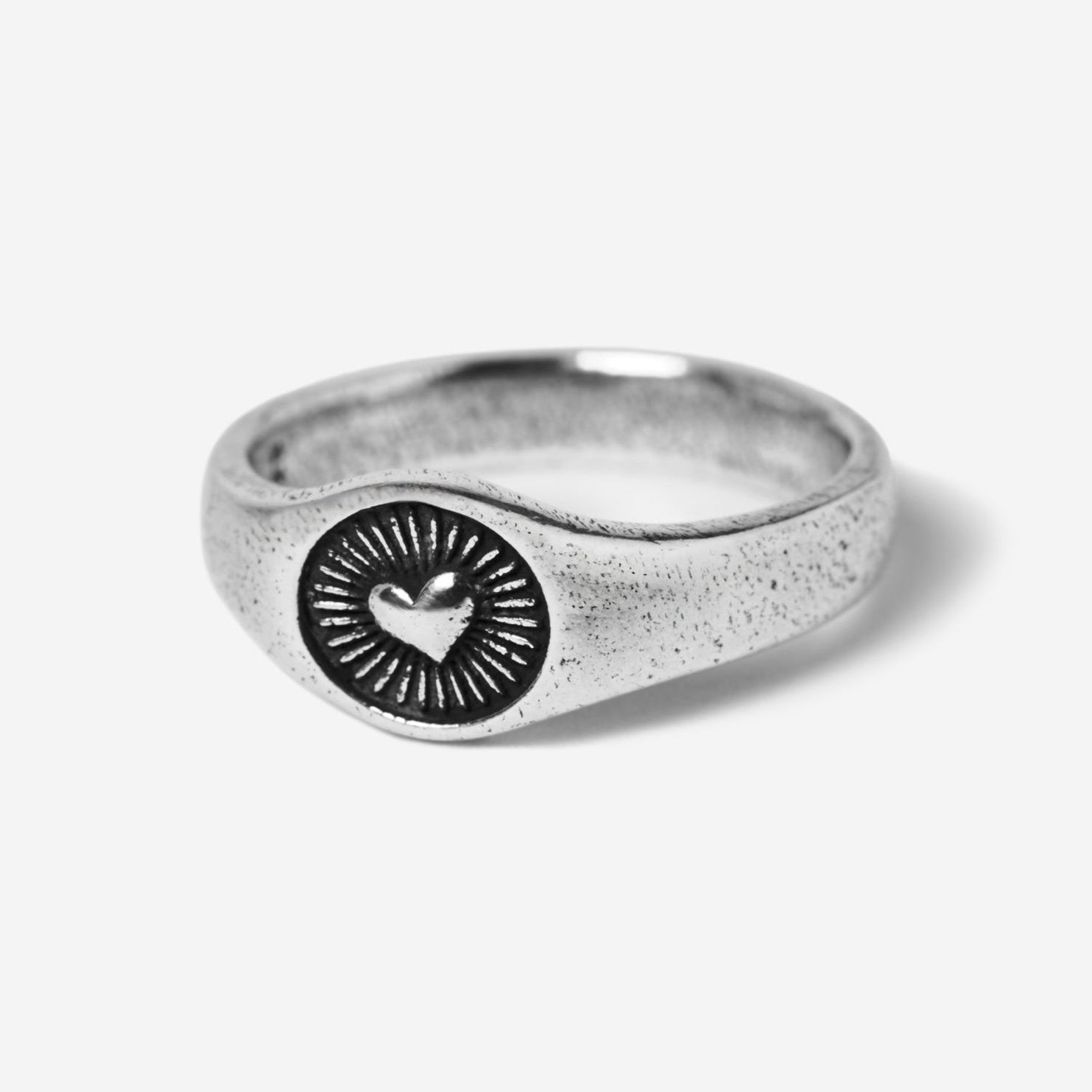 A 45-degree product shot of the sterling silver Vita Ring by Billie Jo. The signet-style ring features a central heart emblem with radiating sunbeam engravings, showcasing its textured, oxidised finish.