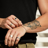 A man with a tiger tattoo wears the Billie Jo ONYX 2.0 RING, a stack of other silver rings, and a bracelet. He adjusts his bracelet, showcasing the men's jewellery.