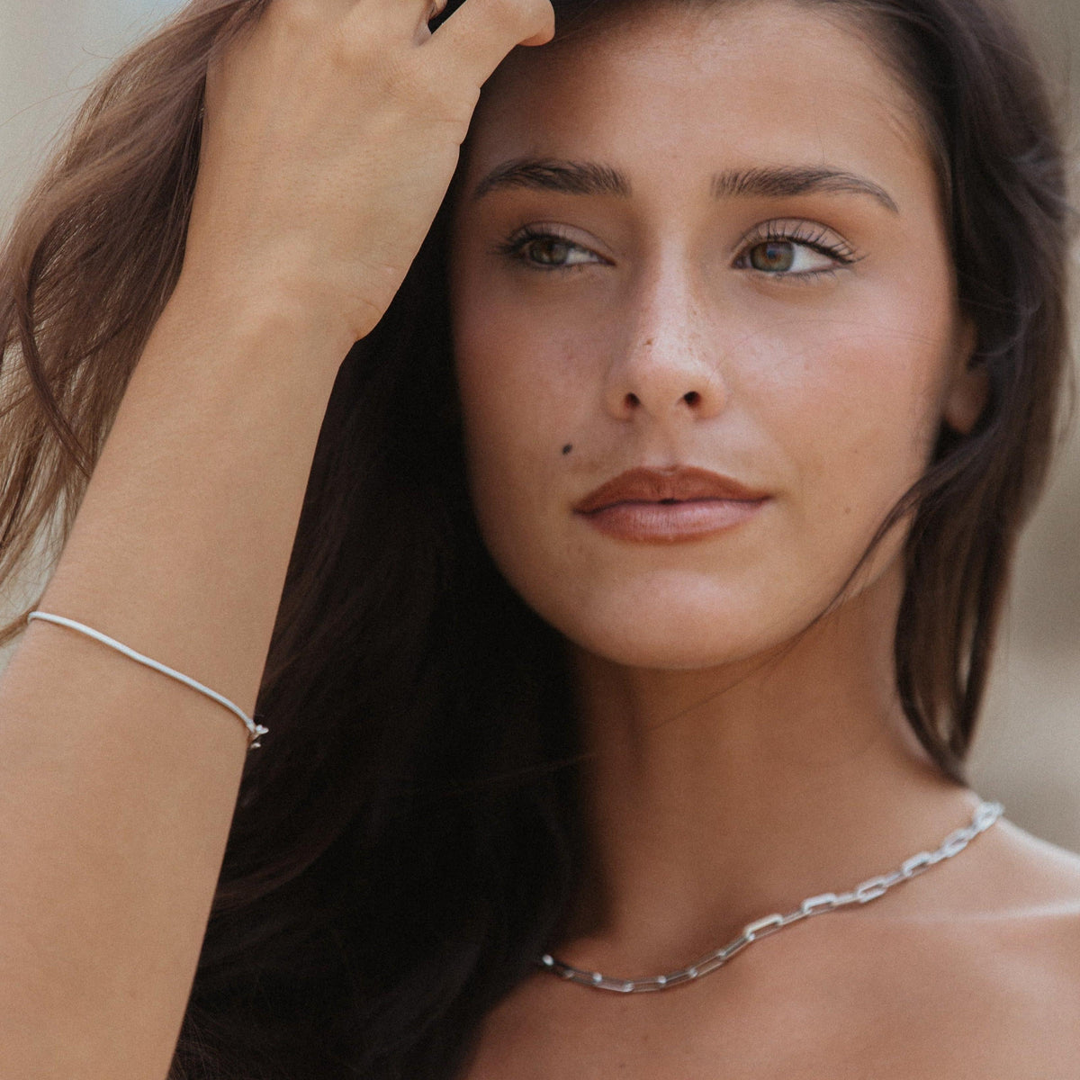 A woman with brunette hair gazes to the side, her hand brushing through her hair, revealing the sterling silver Billie Jo Snake Bracelet on her wrist. This lifestyle shot highlights the bracelet's delicate and versatile design.