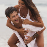 A man gives a woman a piggyback ride on the beach, both smiling and wearing Billie Jo jewellery. He wears the sterling silver Snake Bracelet, adding a touch of modern style to this candid, sun-drenched moment.