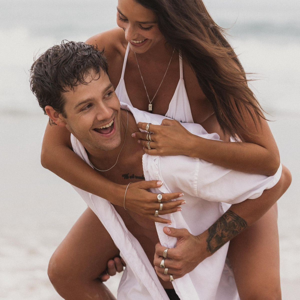 A man gives a woman a piggyback ride on the beach, both smiling and wearing Billie Jo jewellery. He wears the sterling silver Snake Bracelet, adding a touch of modern style to this candid, sun-drenched moment.