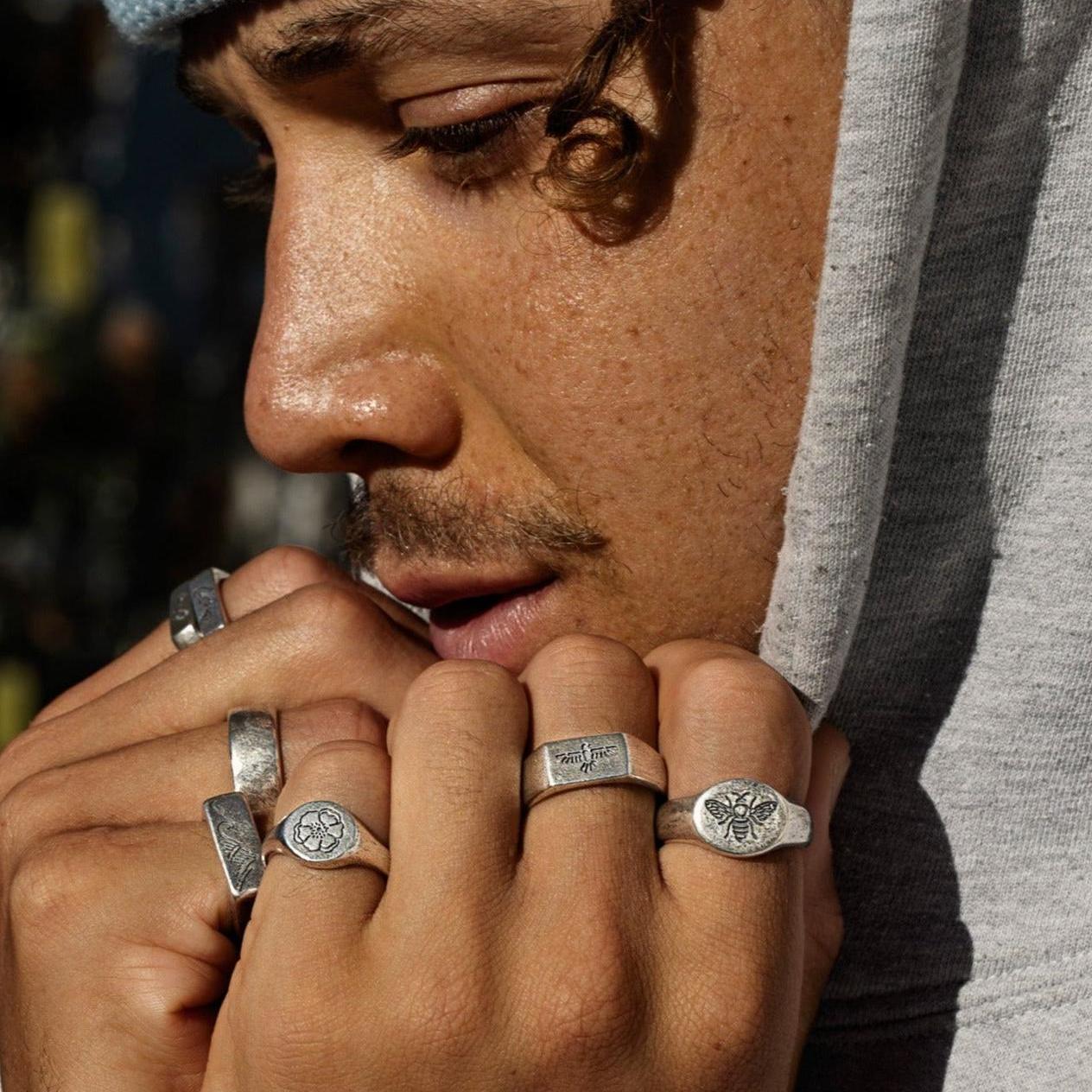 A male model in a grey hoodie with his hands to his face, showcasing a stack of men's rings including the BEE THERE RING by Billie Jo.