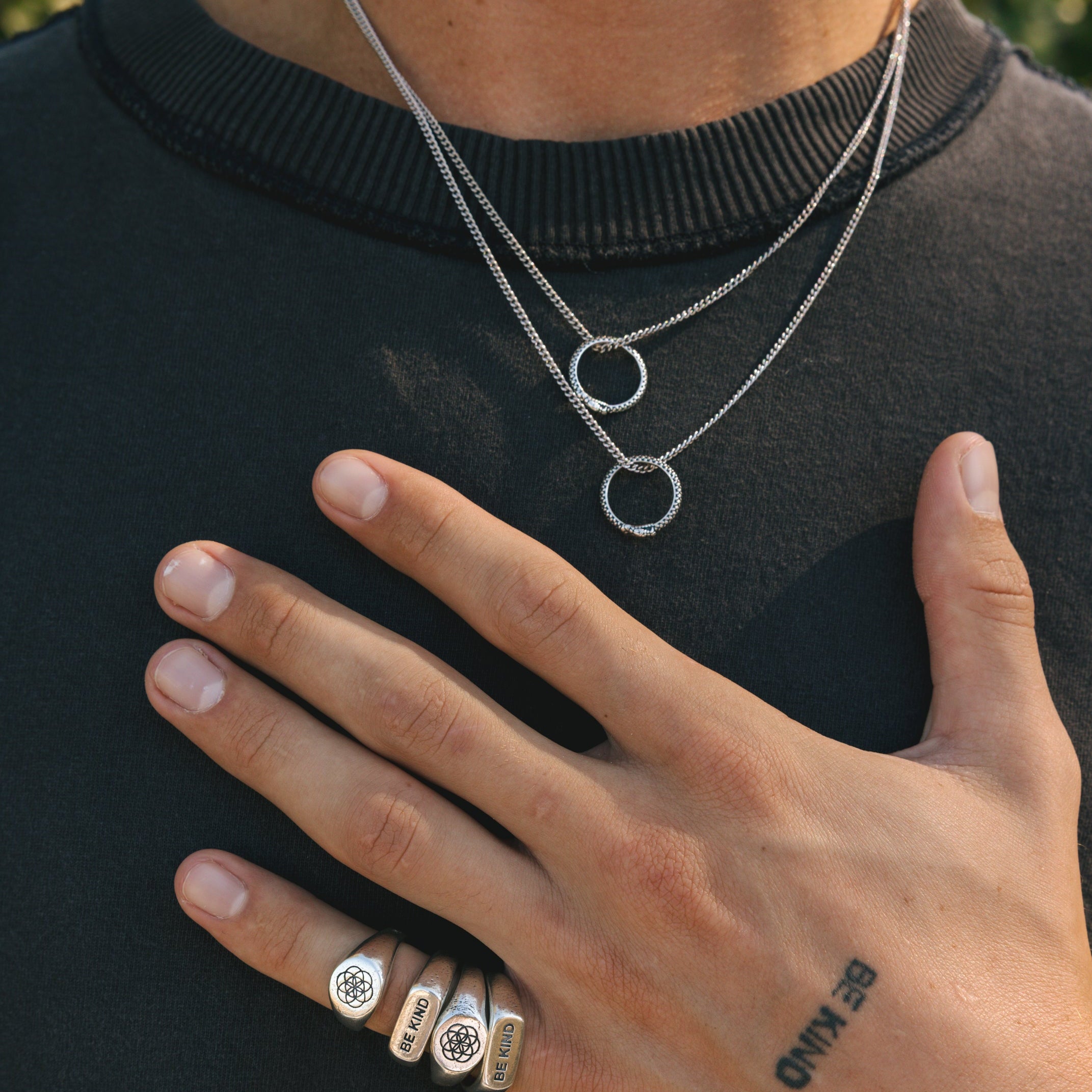 A man wearing two layered Billie Jo Ouroboros Pendants and multiple silver rings against a black tee. A modern, masculine look for Australian men's jewellery.