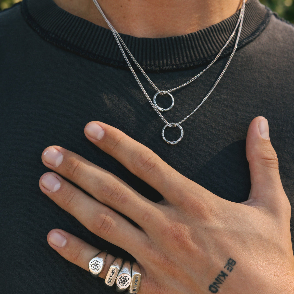 A man wearing two layered Billie Jo Ouroboros Pendants and multiple silver rings against a black tee. A modern, masculine look for Australian men's jewellery.