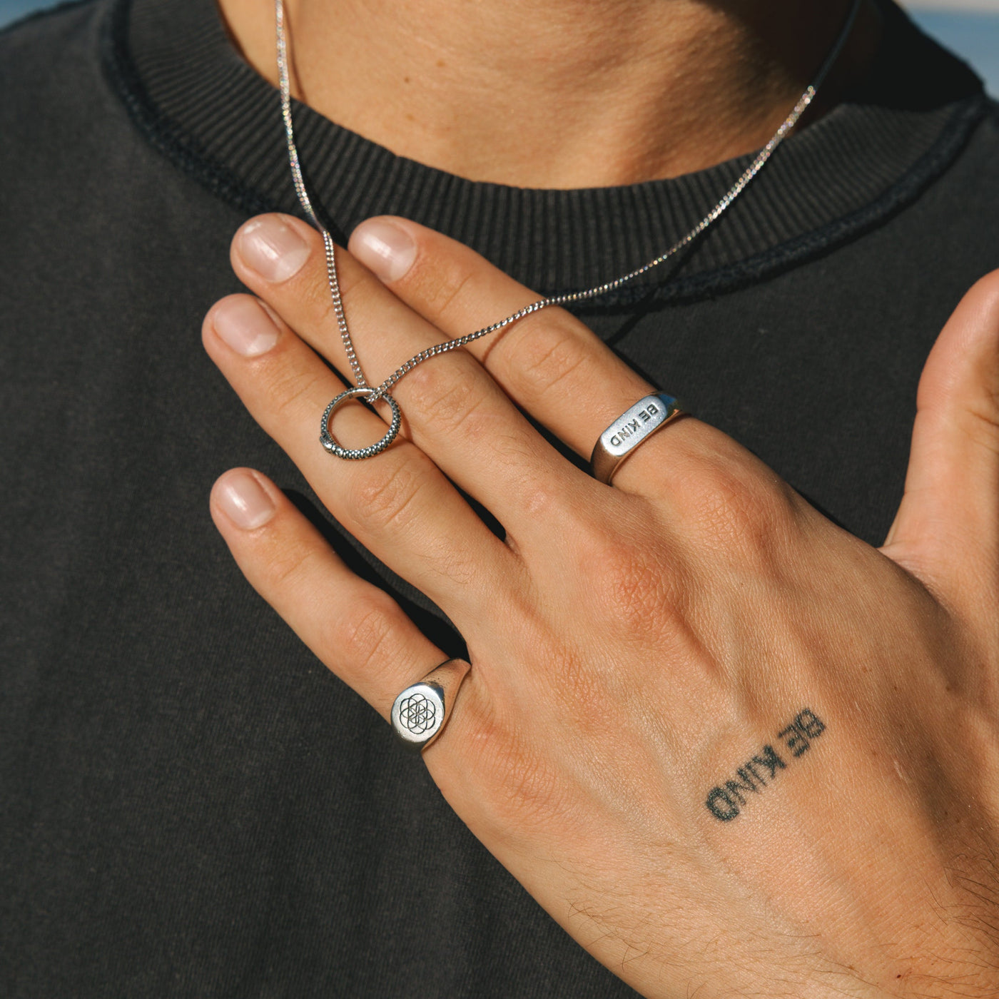 A man wearing the Billie Jo UNITY RING and a sterling silver necklace, with his tattooed hand resting on his chest against a black t-shirt.