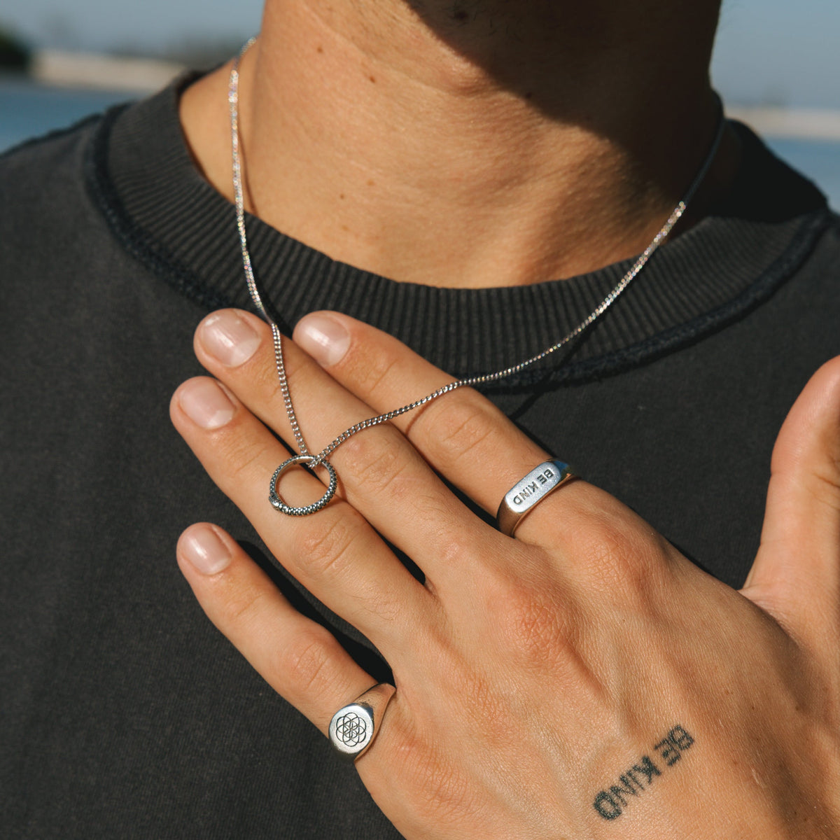 A man wearing the sterling silver Ouroboros Pendant from Billie Jo, styled with a black tee. His hand, showing 'BE KIND' tattoo, also wears silver rings.