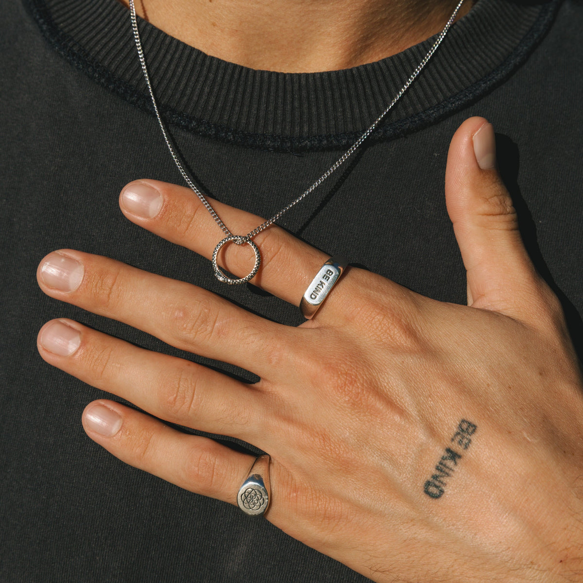 A man wearing the sterling silver BE KIND ring from Billie Jo on his index finger, styled with a matching tattoo and silver necklace for a modern look.