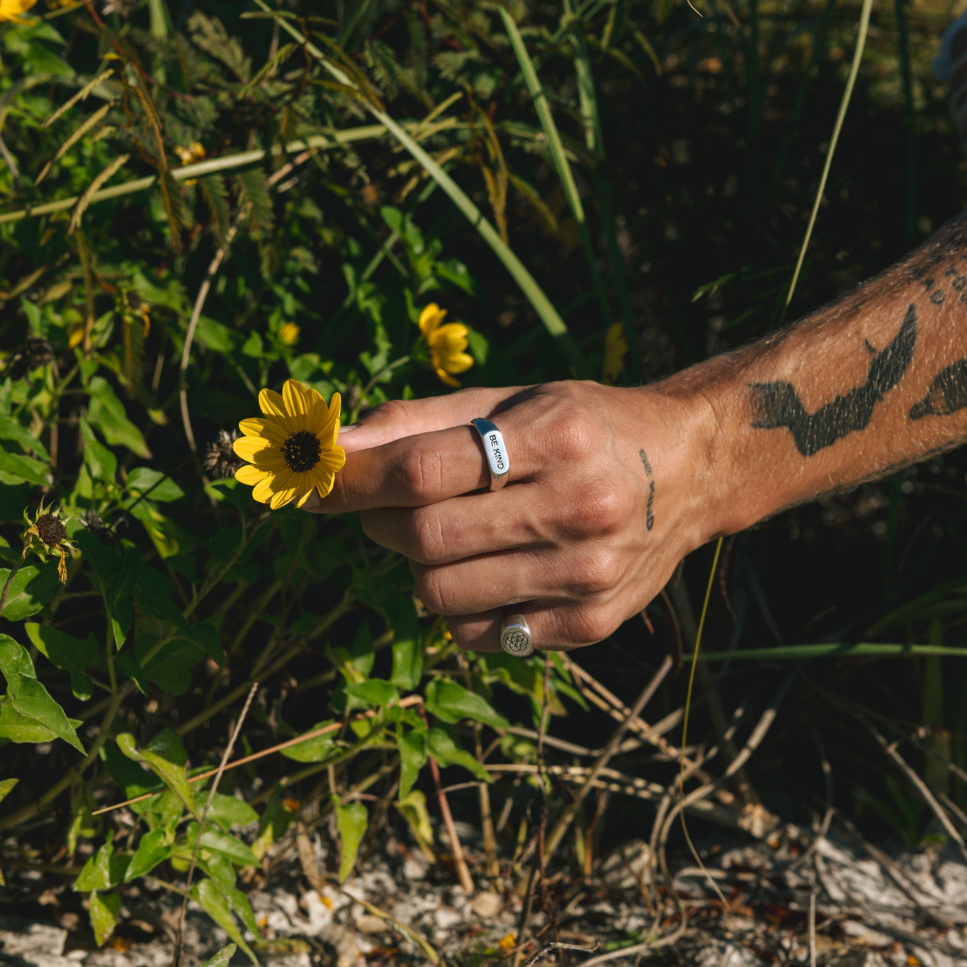 An artistic shot of a man's hand holding a yellow flower, wearing the BE KIND ring by Billie Jo. A blend of nature and modern men's jewellery.