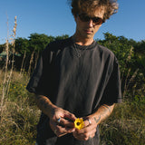A man wearing the Billie Jo Ouroboros Pendant in a sunny, coastal field, holding a yellow wildflower. An aesthetic shot for this men's jewellery piece.