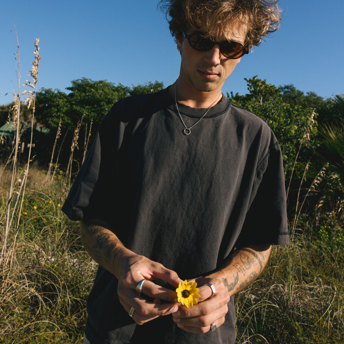 A man wearing the Billie Jo Ouroboros Pendant in a sunny, coastal field, holding a yellow wildflower. An aesthetic shot for this men's jewellery piece.