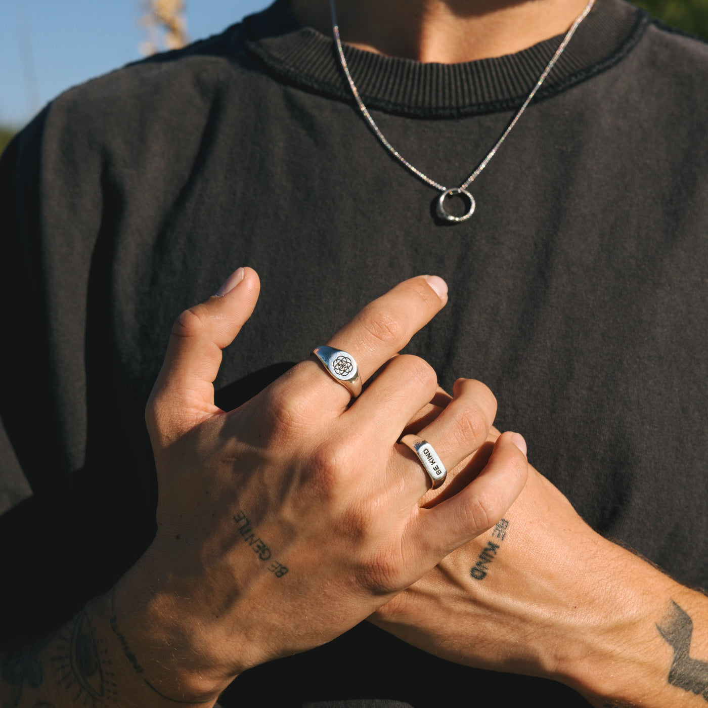 A man's hands with the Billie Jo UNITY RING and another silver band, styled with a silver necklace against a black t-shirt in a sunny outdoor setting.