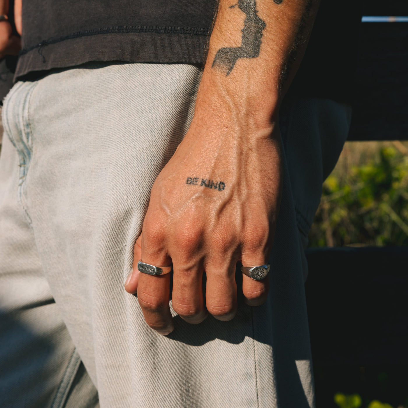 Detailed view of the BE KIND ring on a man's hand, showing the texture of the silver and how it sits alongside other rings and tattoos.