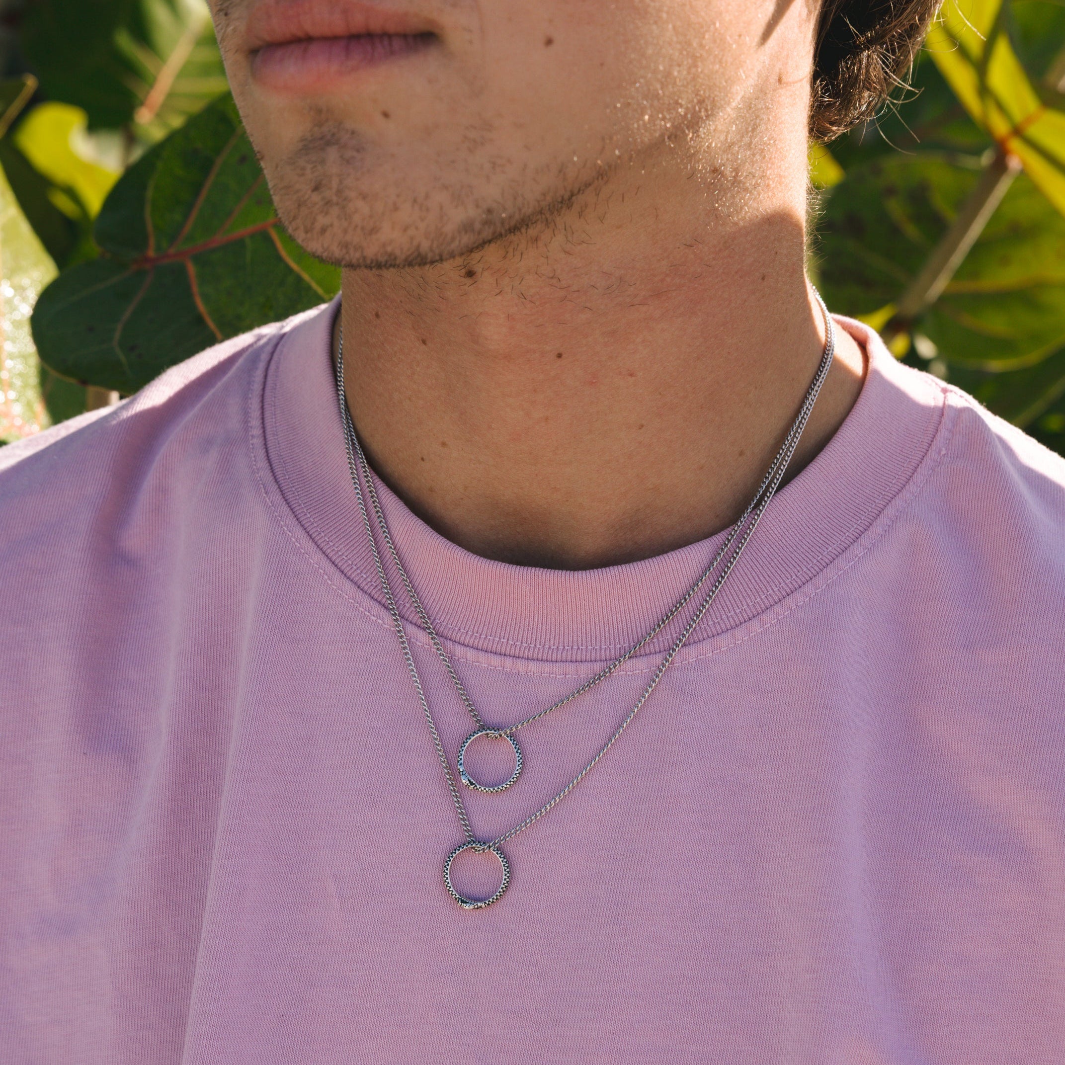 Layered styling of the Billie Jo Ouroboros Pendant. A man wears two of the sterling silver necklaces with a pink tee for a modern, editorial look.