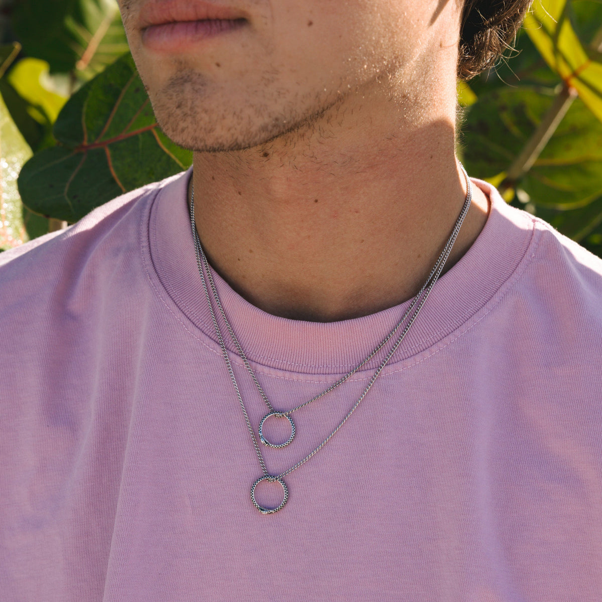 Layered styling of the Billie Jo Ouroboros Pendant. A man wears two of the sterling silver necklaces with a pink tee for a modern, editorial look.