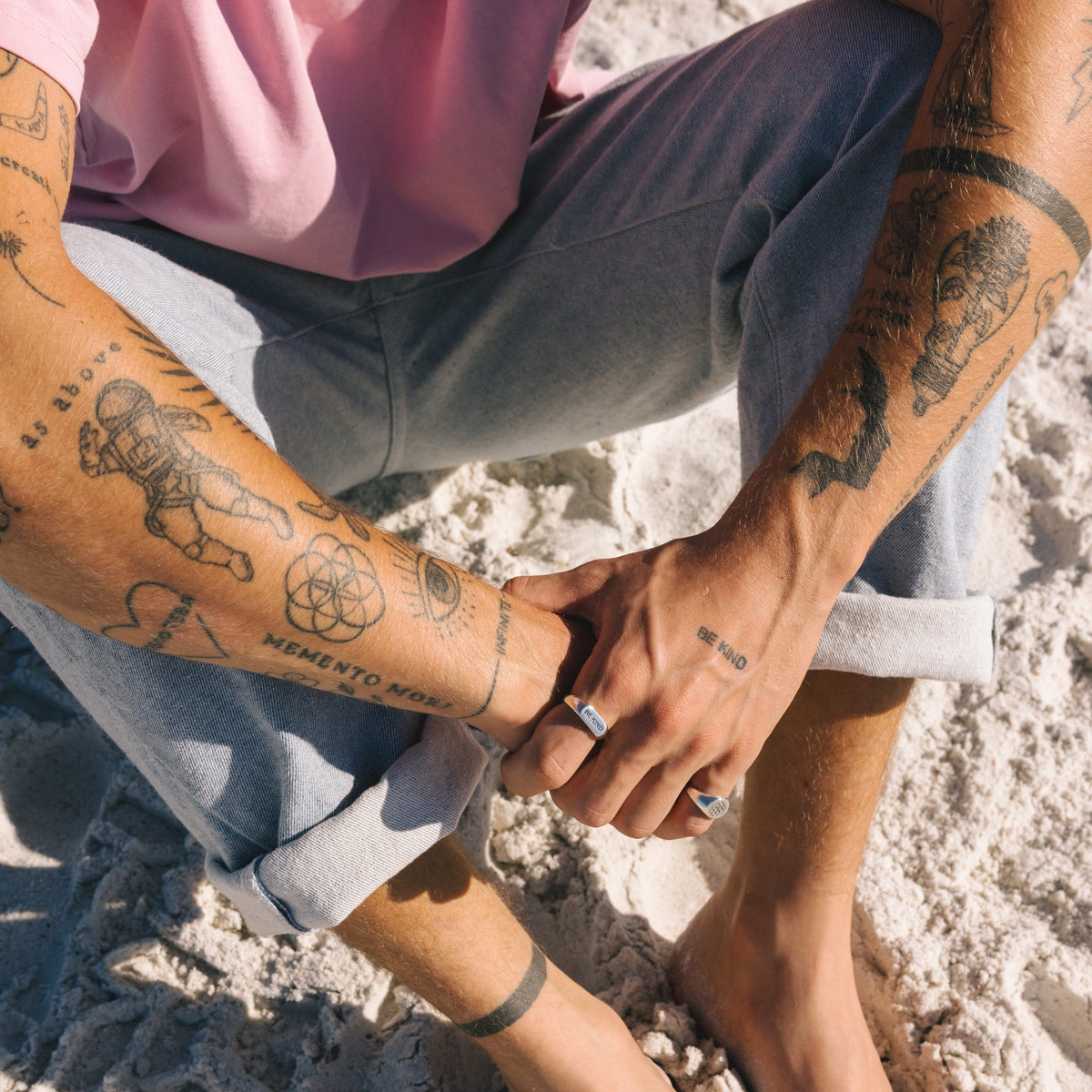 A full-body shot of a man sitting on the beach, wearing the BE KIND ring and other Billie Jo jewellery, embodying a laid-back, Australian style.