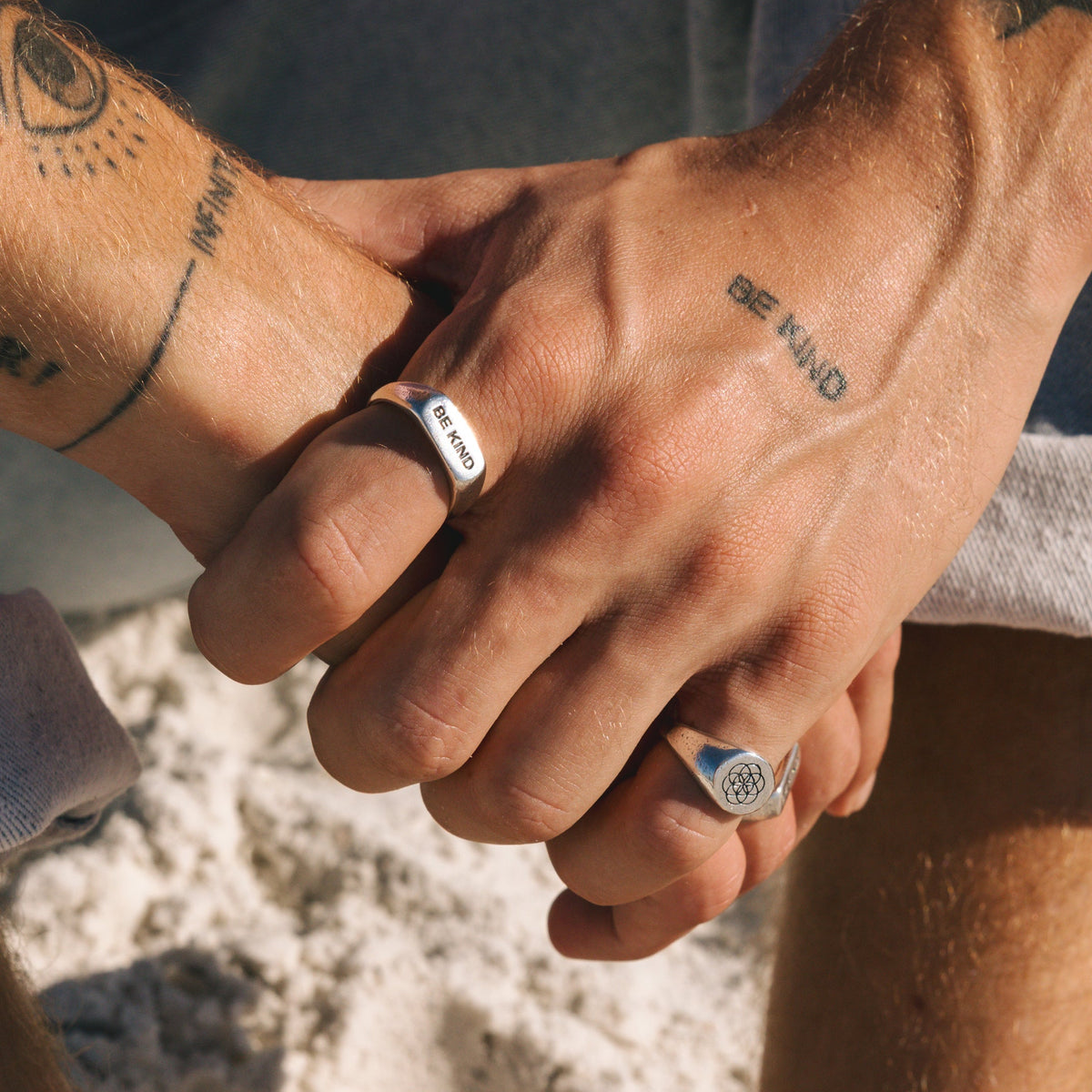 A close-up of two men holding hands on a sandy beach, one wearing the Billie Jo UNITY RING and another silver band, showcasing men's jewellery.