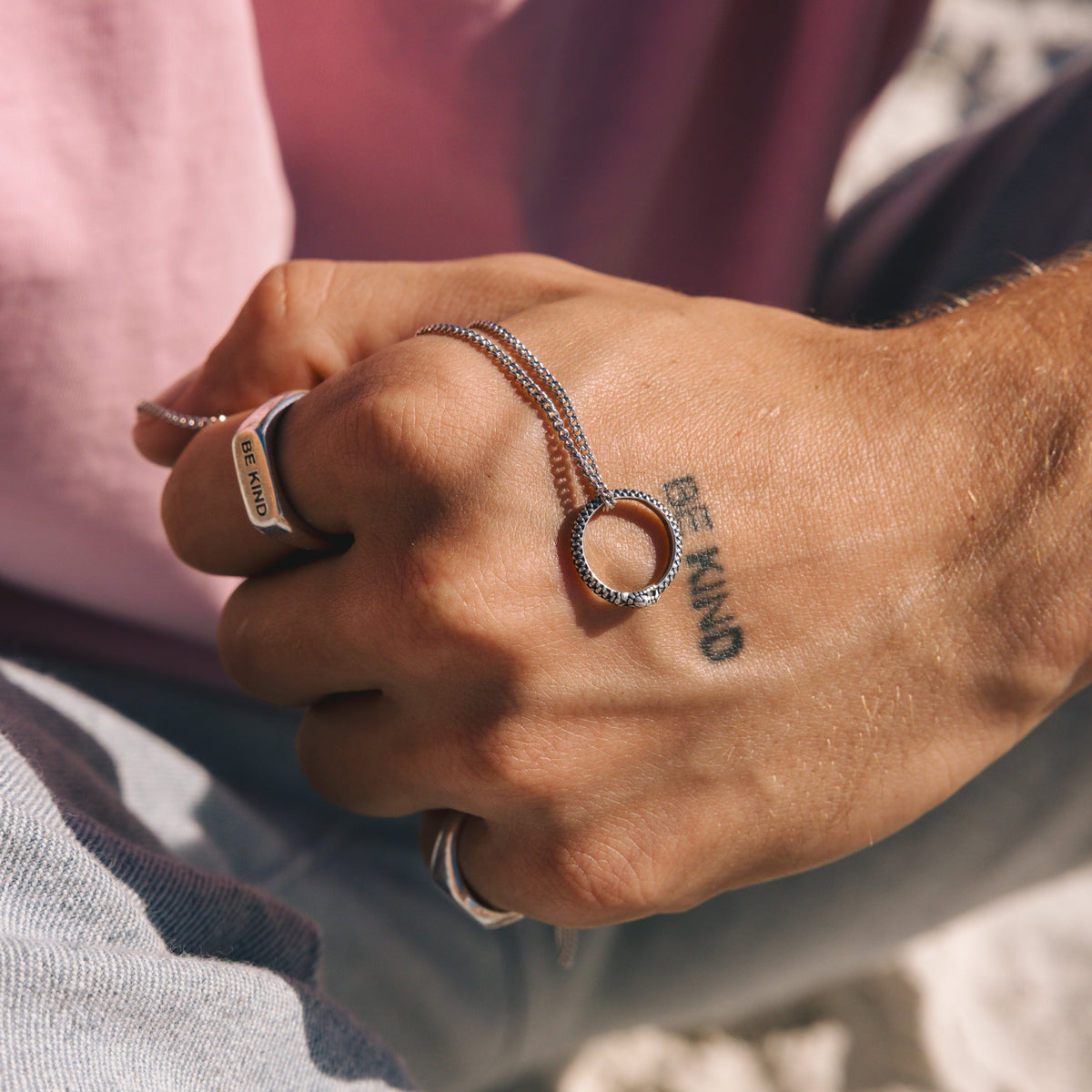 Close-up on a man's hand holding the Ouroboros Pendant, with a 'BE KIND' tattoo and silver rings. Showcasing Billie Jo's men's jewellery styling.