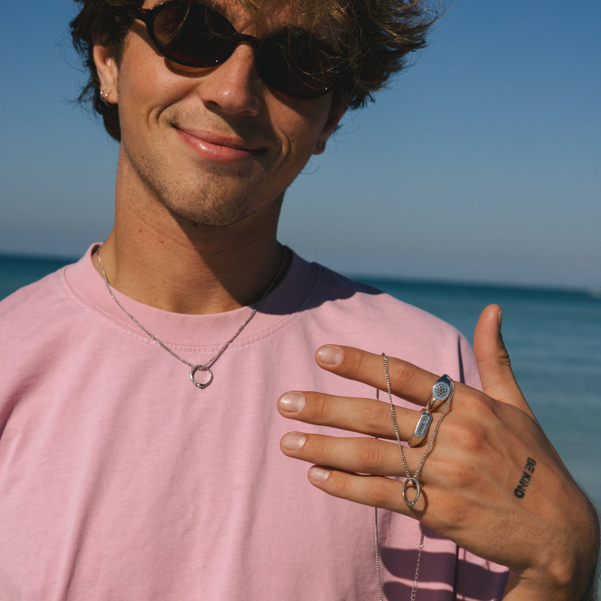 A smiling man on a sunny Australian beach wearing a pink t-shirt, sunglasses, and Billie Jo's sterling silver UNITY RING and necklace.