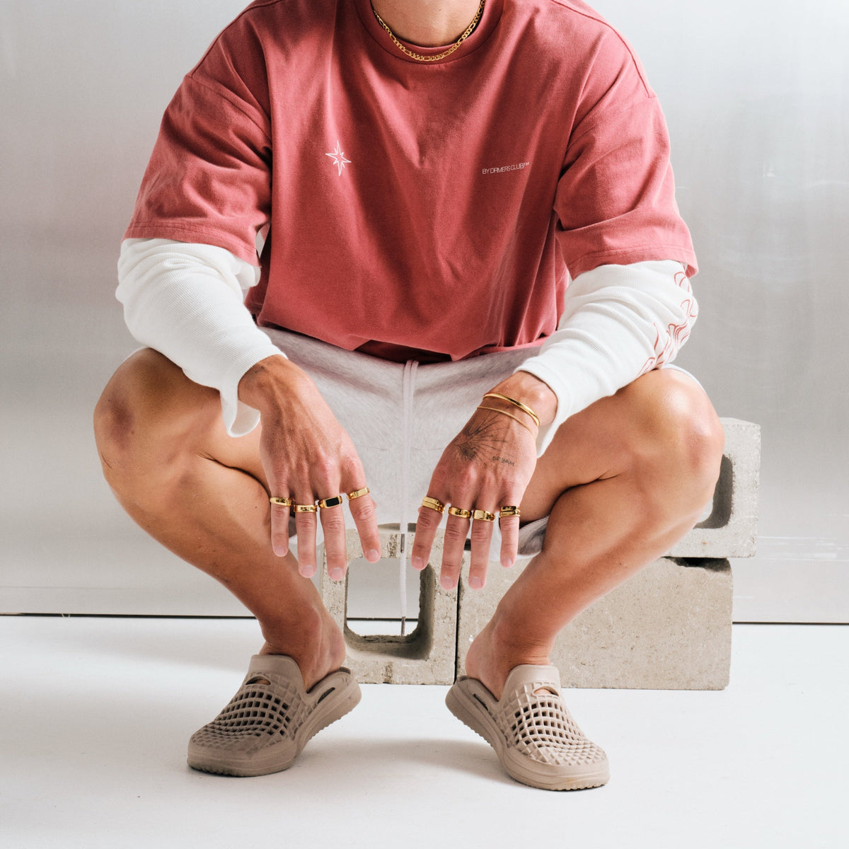 Man wearing gold stainless rings, crouched in front of concrete blocks, with a gold chain necklace, gold bracelets