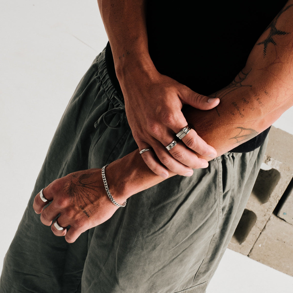 Man wearing silver finish stainless rings, arms crossed, concrete block behind, with a steel Cuban bracelet