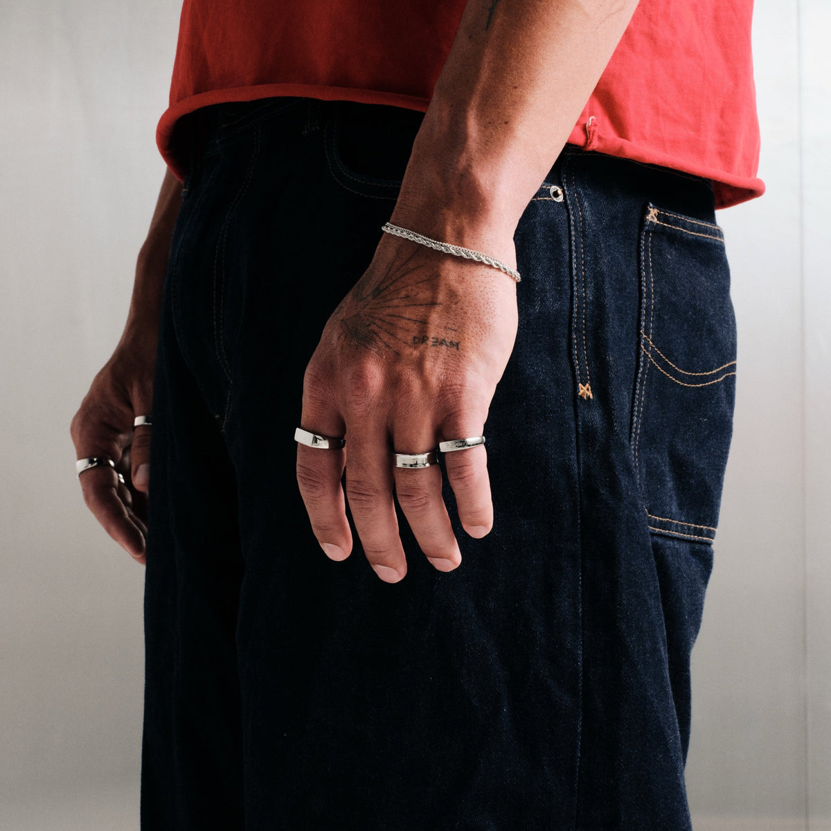 Man wearing silver finish stainless rings, hand by his side, with a steel rope bracelet, red tee, dark jeans