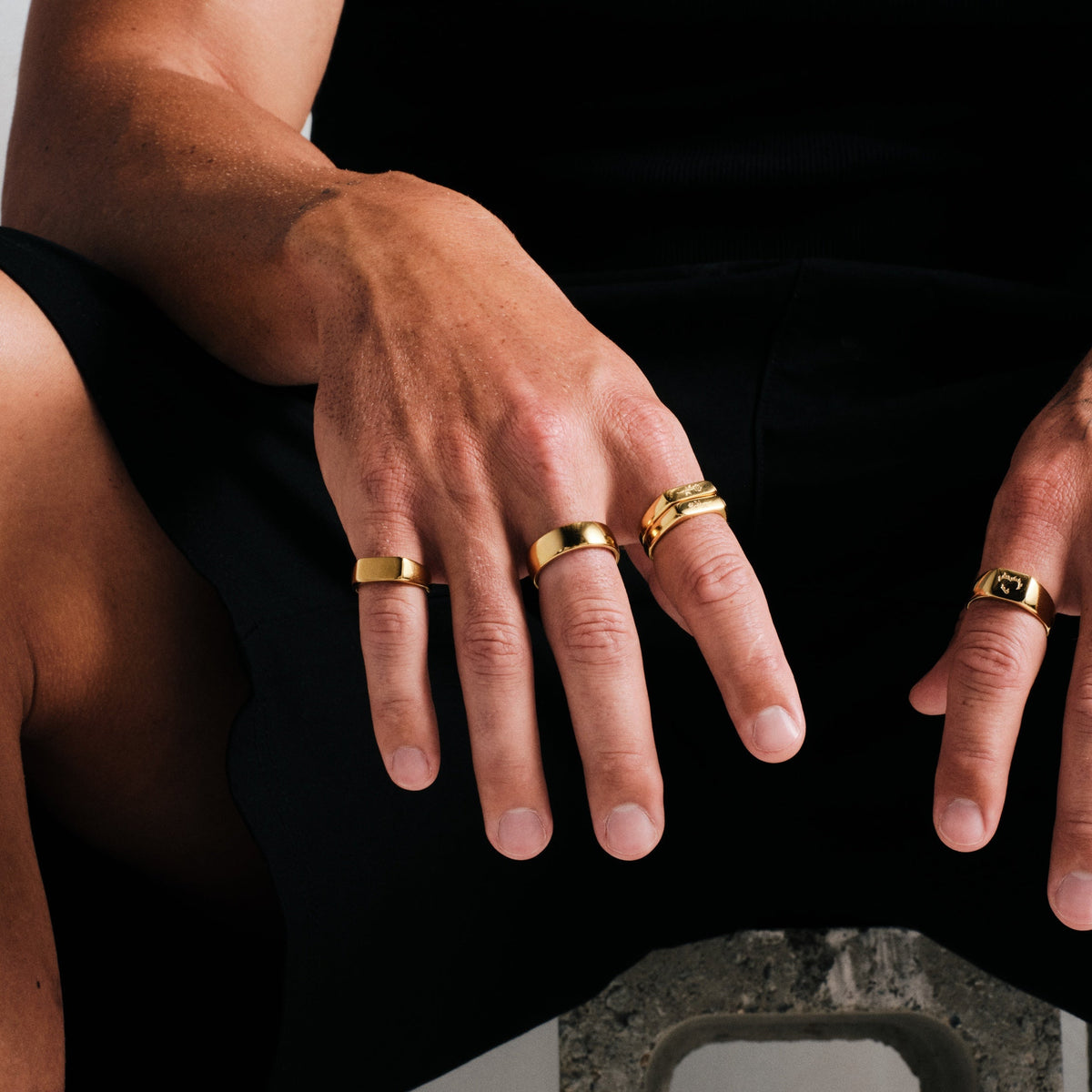 Men's Gold Genesis Ring in 18k gold infused stainless steel, in a stack, seated on concrete block, hands on knees
