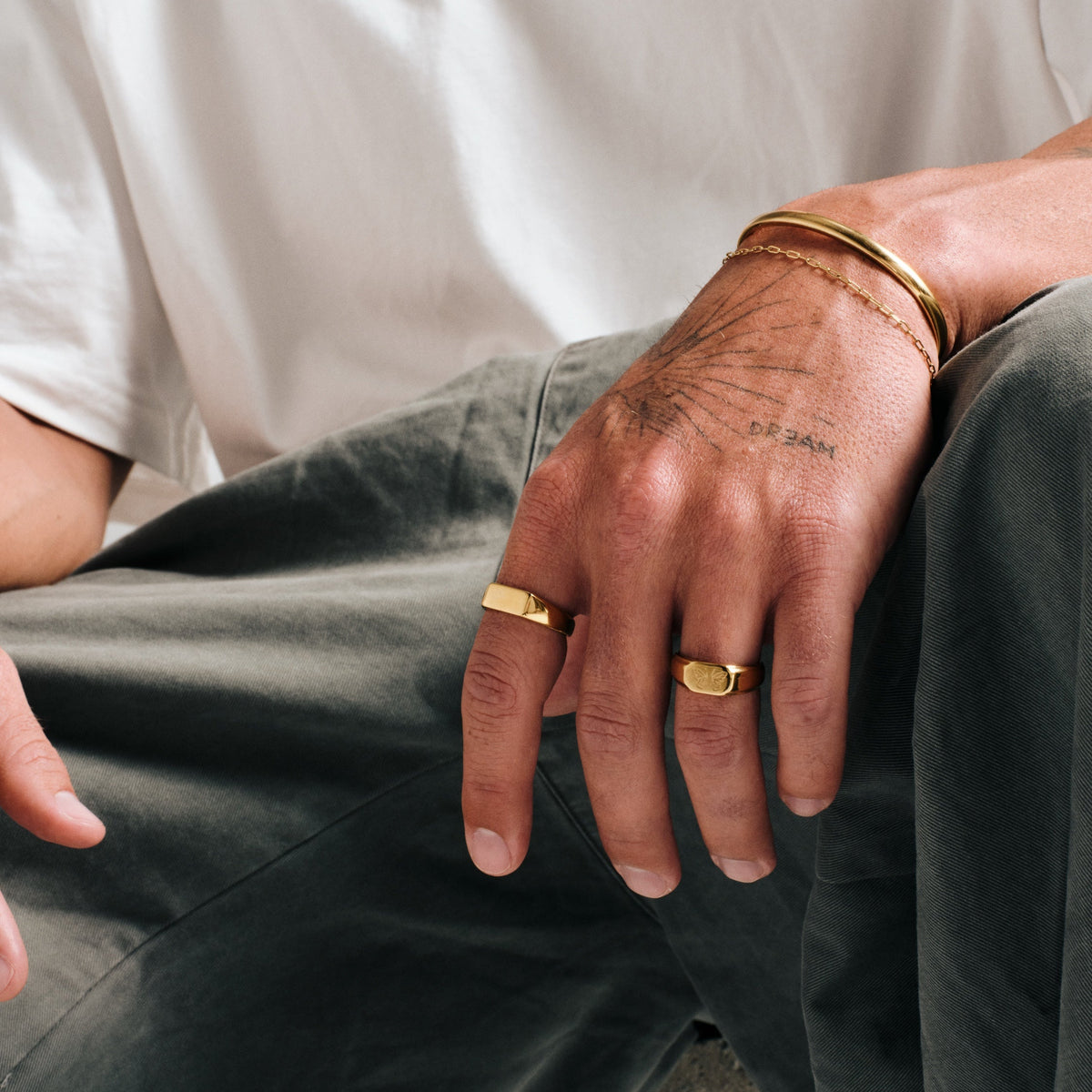 Men's Gold Genesis Ring in 18k gold infused stainless steel, worn on his left index finger, seated, hand resting on knee