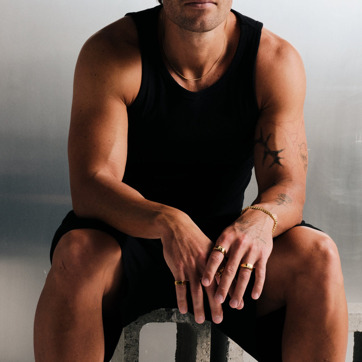 Man wearing gold stainless rings, seated on concrete block, hands clasped, black singlet, black shorts