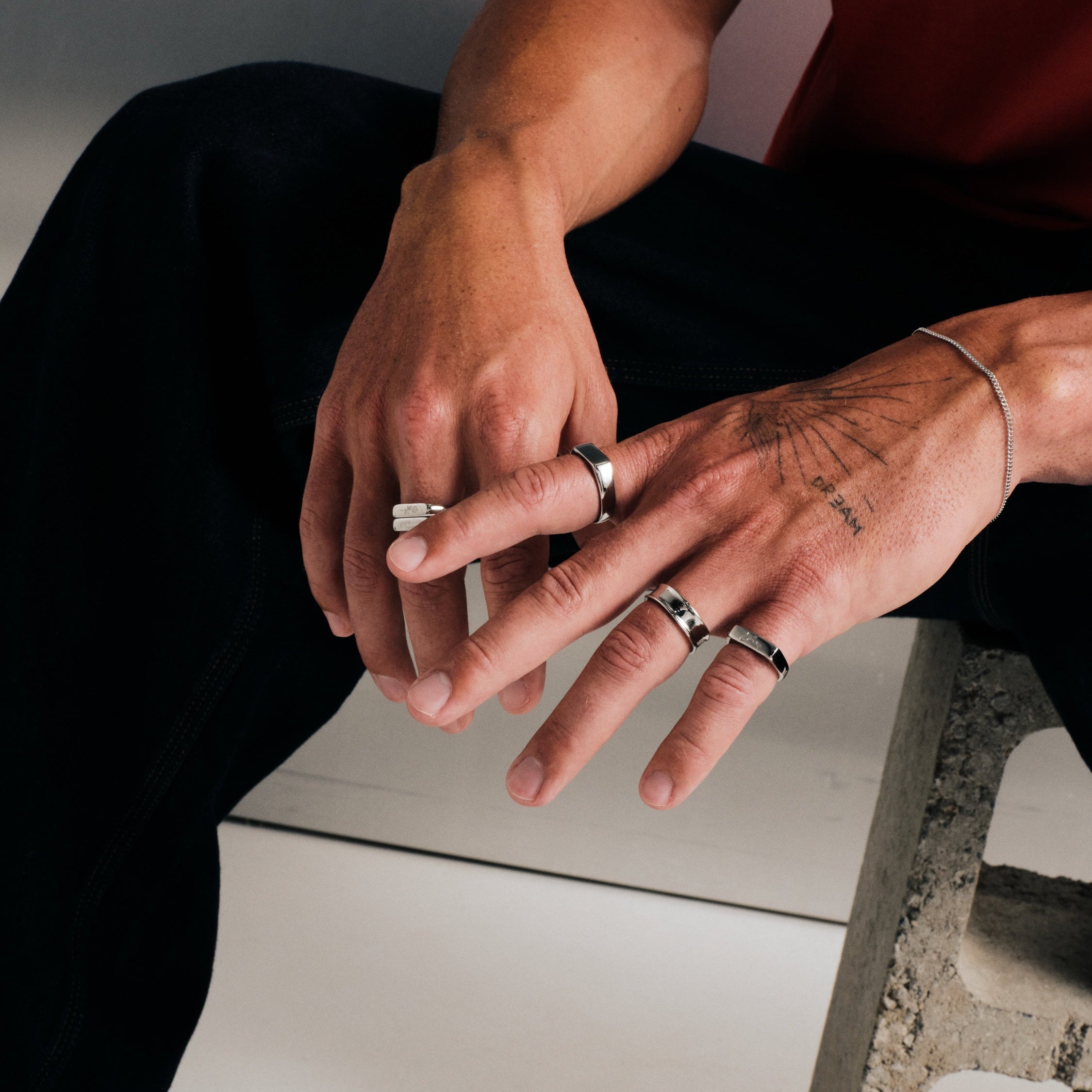 Man wearing silver finish stainless rings, seated, one hand over the other, with a slim chain bracelet, red tee, black pants