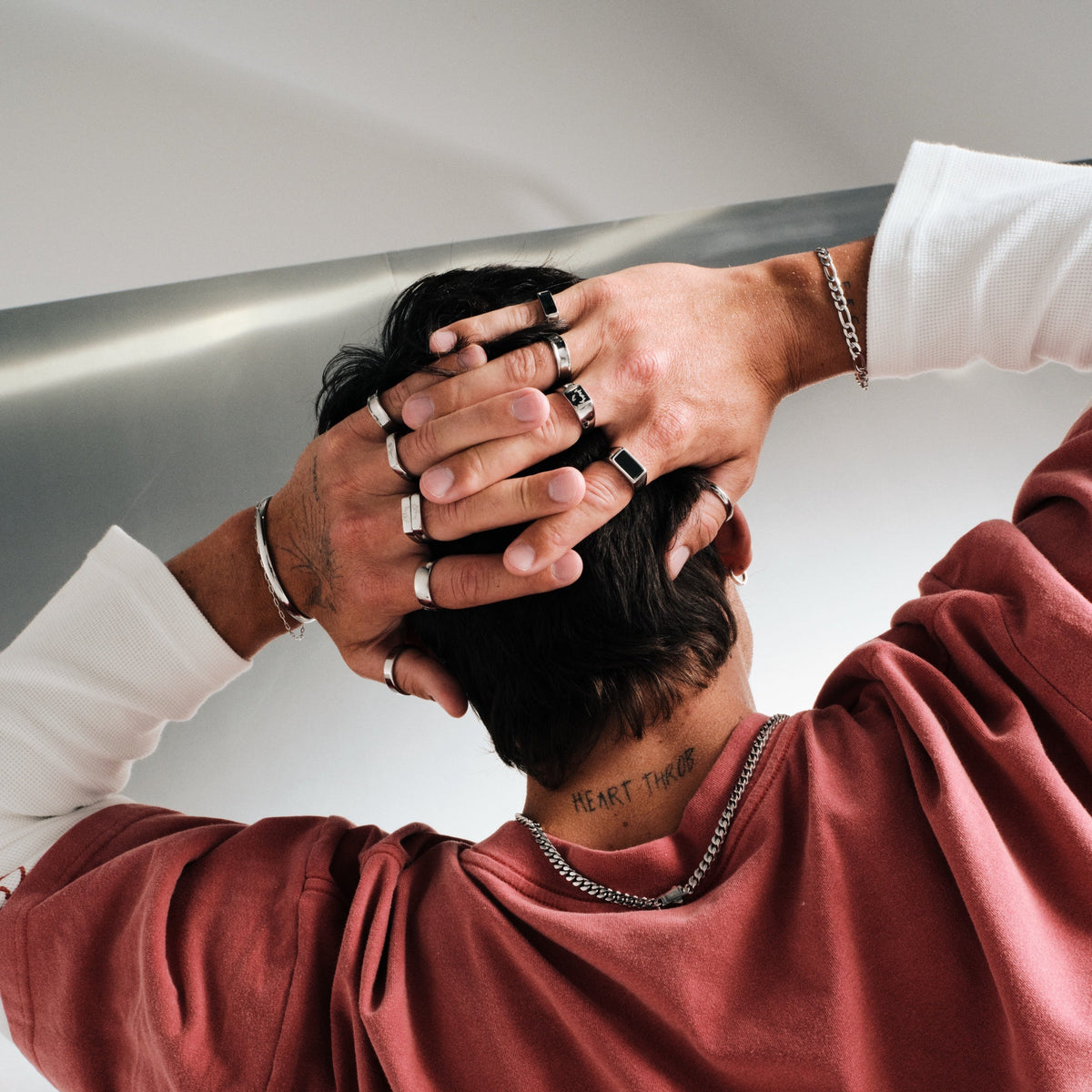 Man wearing silver finish stainless rings, hands behind his head, back to camera, red tee over white long sleeve