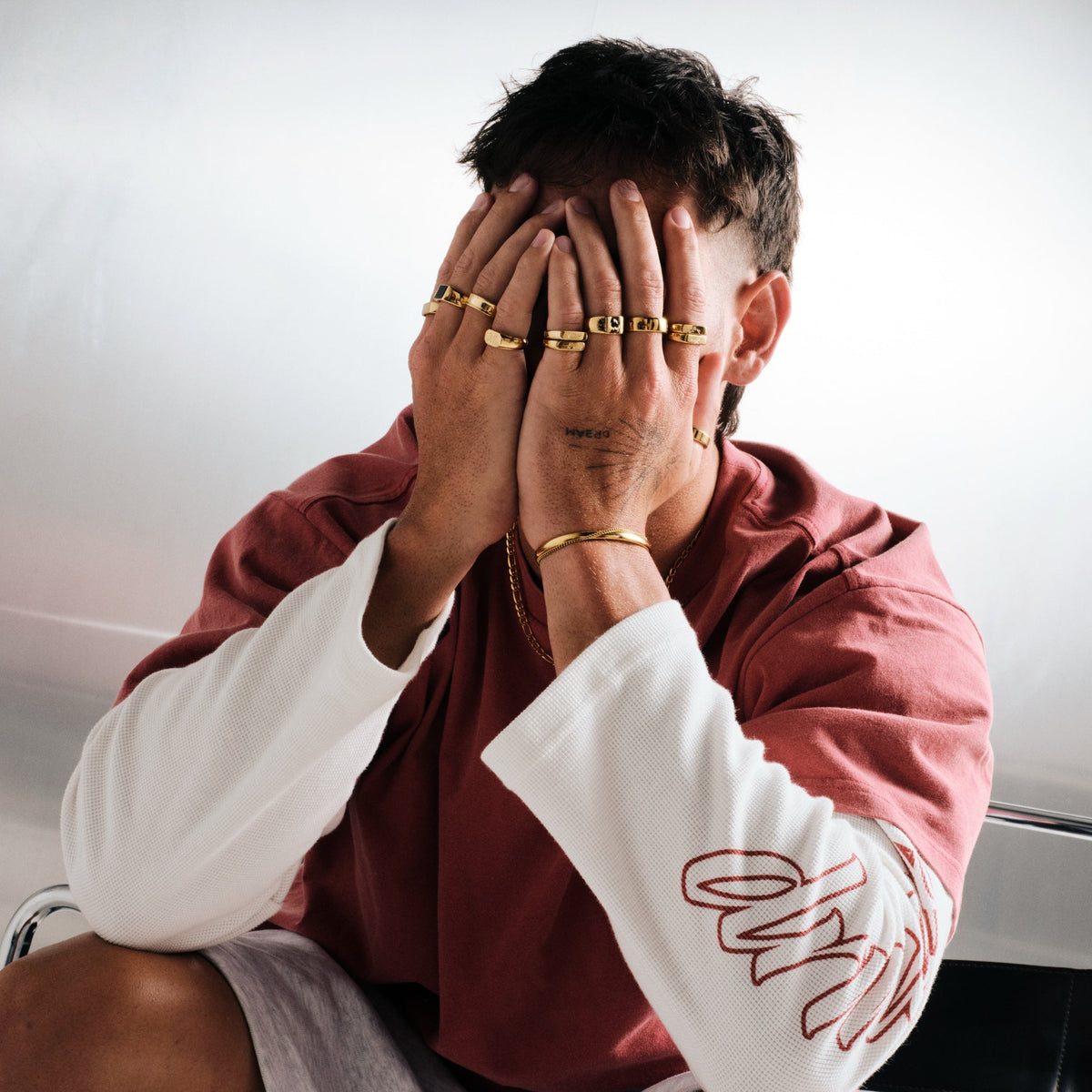 Man wearing gold stainless rings, seated, hands covering his face, with a gold chain necklace, gold cuff on left wrist