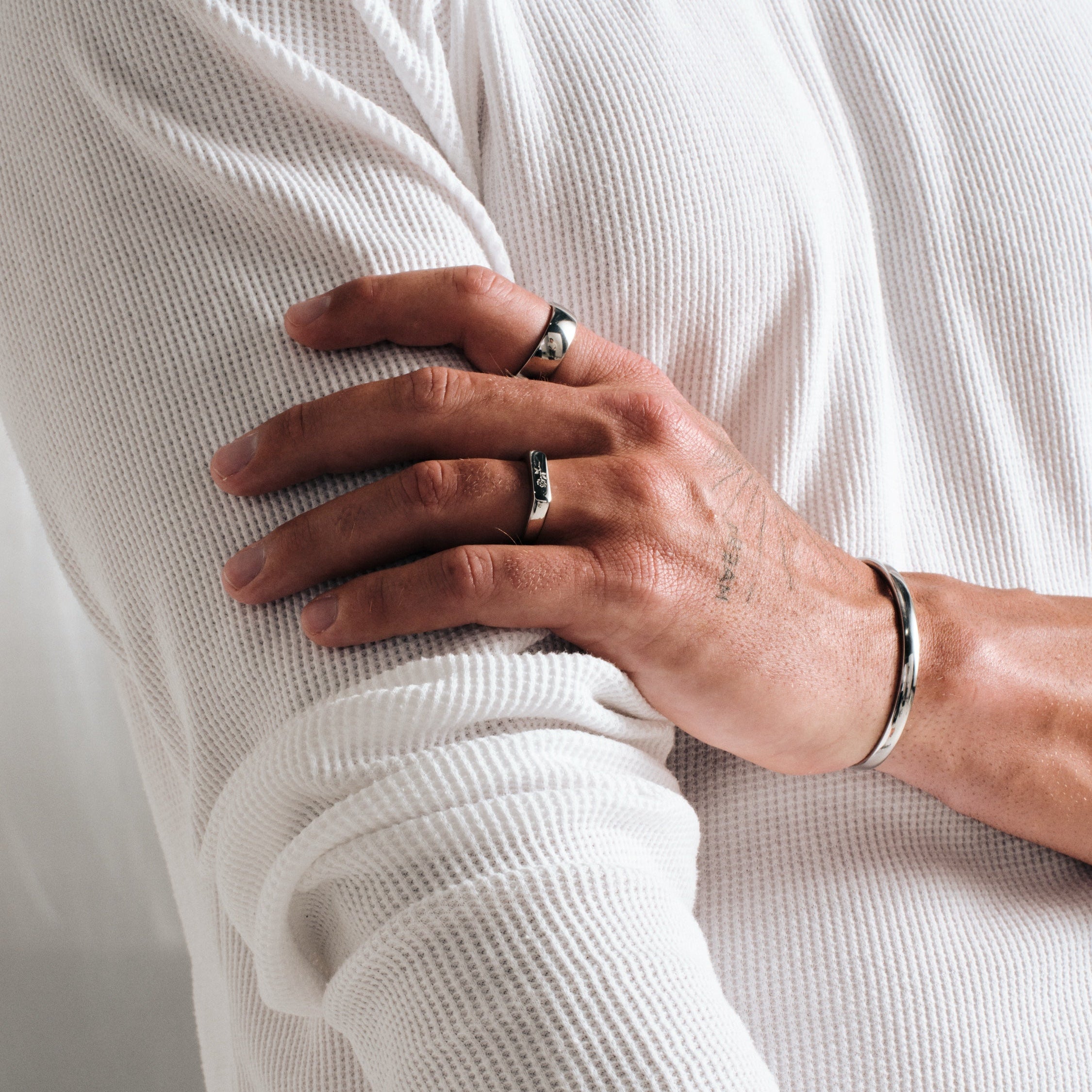 Man wearing silver finish stainless rings, hand holding his upper arm, with a steel Drifter Cuff on wrist