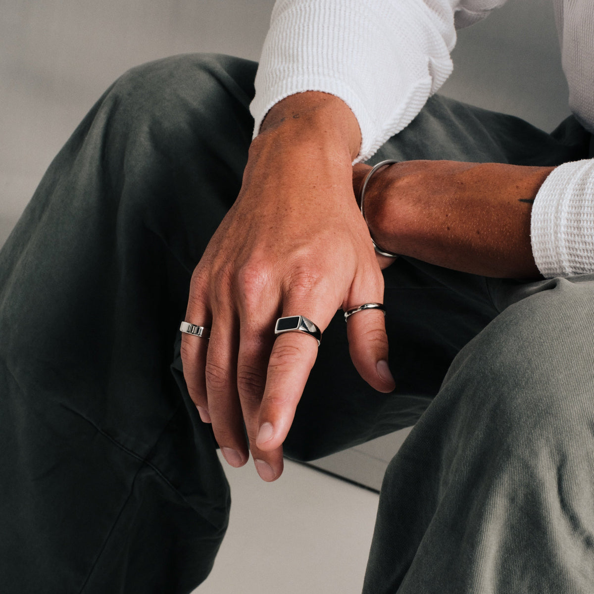 Man wearing silver finish stainless rings, seated, hand over his knee, with a bangle cuff on other wrist