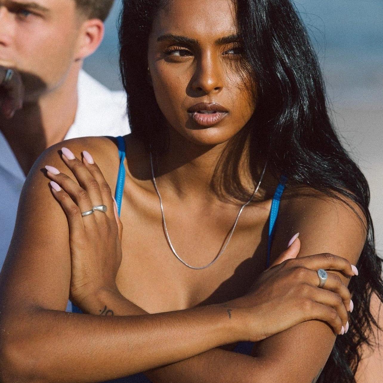 A woman wearing the sterling silver Billie Jo Snake Necklace in a beach setting, her arms crossed. The necklace provides a simple, elegant accent to her blue swimsuit.