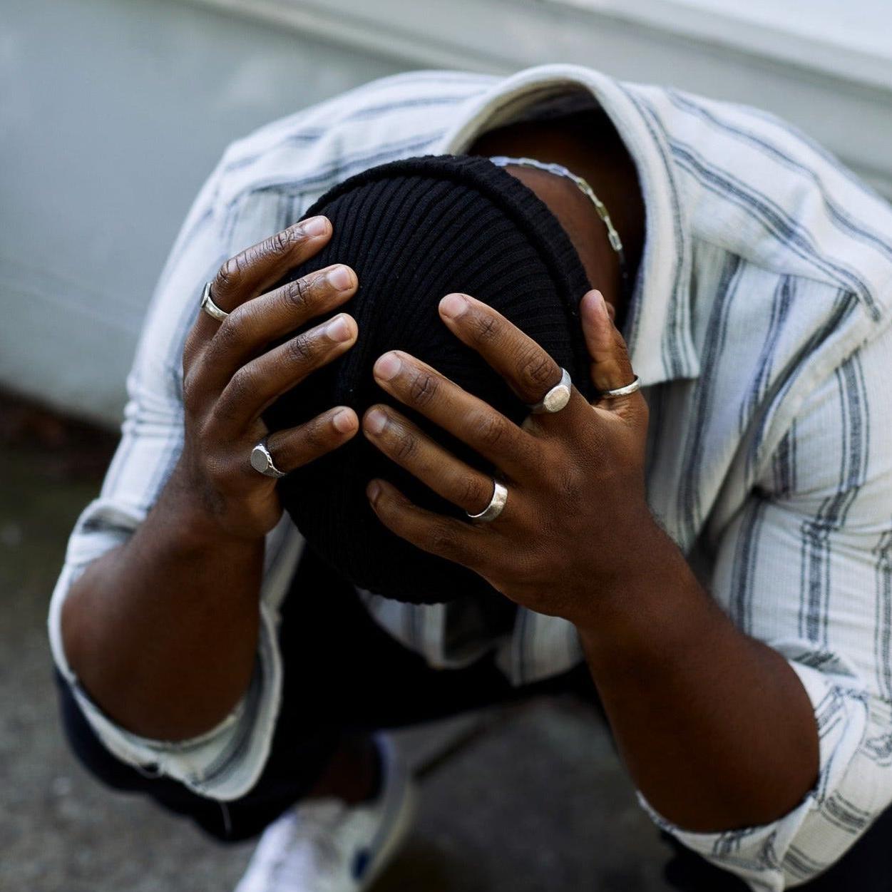 A male model in a striped shirt and beanie, holding his head in his hands, showcasing the Billie Jo Canvas Ring and other men's jewellery.