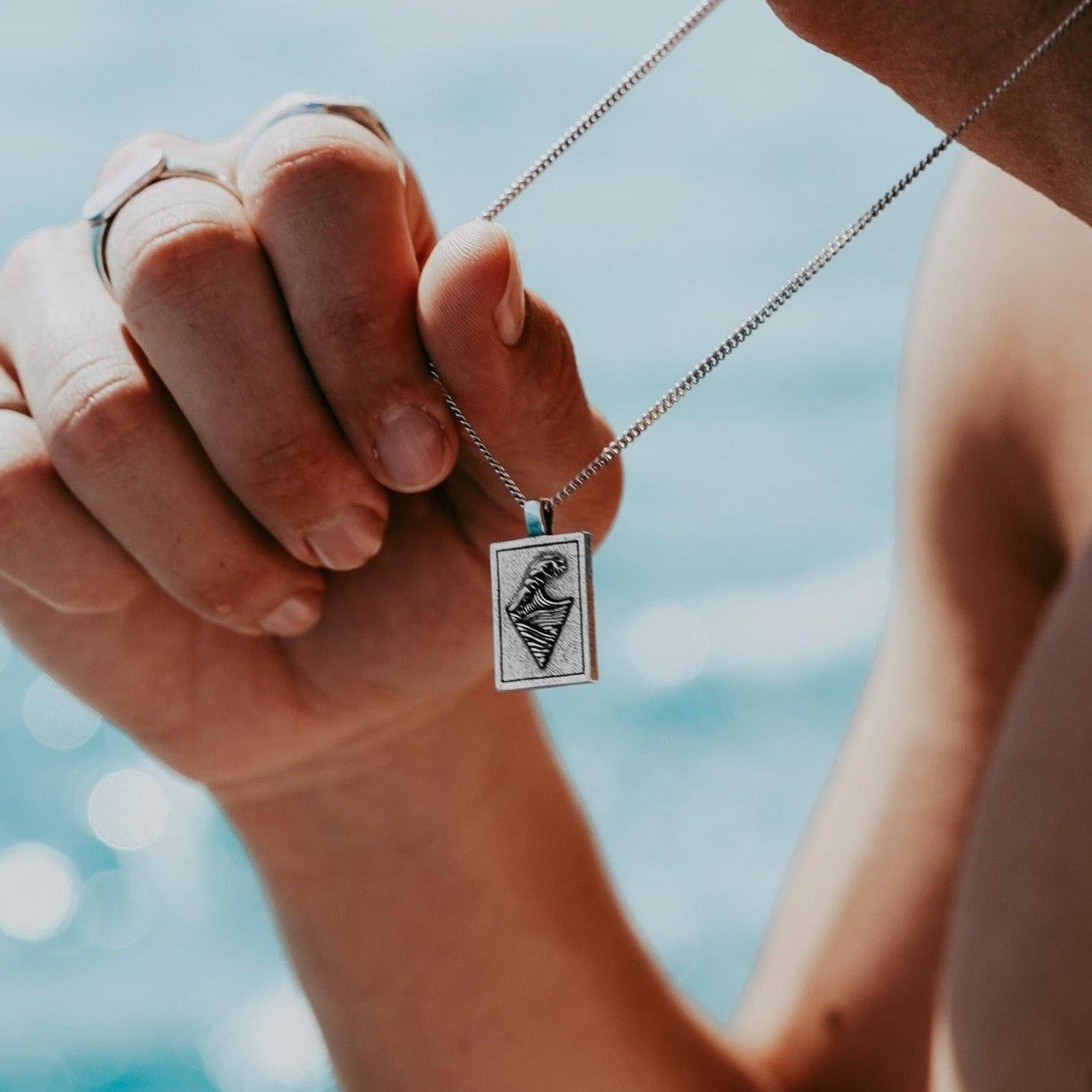 The Burleigh Pendant by Billie Jo, held in a man's hand to show the scale of the rectangular sterling silver pendant with its unique wave design.