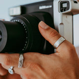 A lifestyle image showing a man's hand holding a camera, wearing the Billie Jo Canvas Ring. This highlights the ring's creative and modern aesthetic.