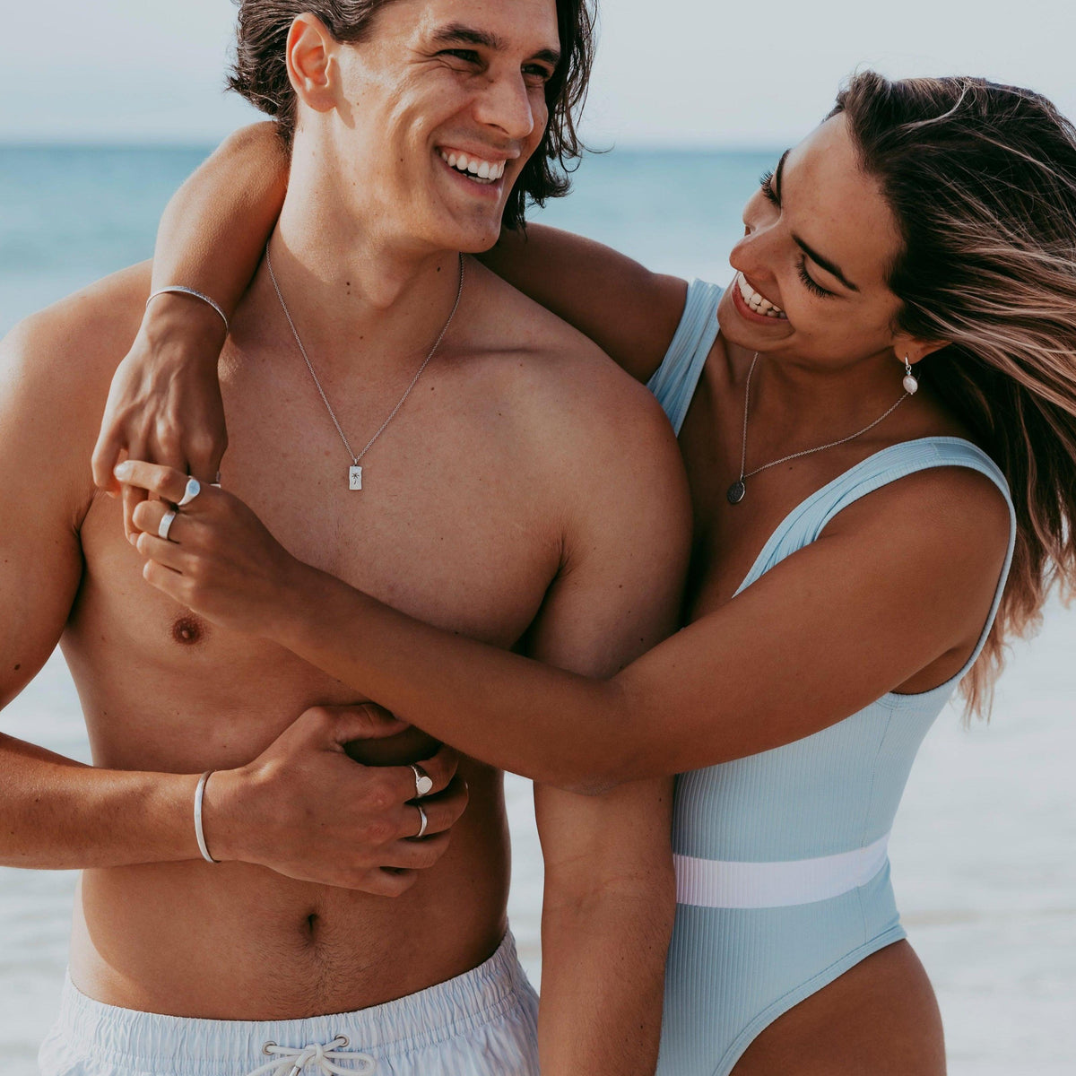 A happy couple on the beach, the man wearing the sterling silver Paradiso Pendant from Billie Jo, showcasing a relaxed, coastal lifestyle.