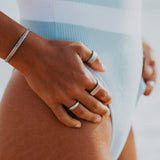 A close-up of a man's hand resting on his hip, adorned with a variety of sterling silver Billie Jo Stacker Rings and a textured bracelet, conveying a relaxed, beachside aesthetic.
