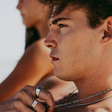 A man with a thoughtful expression adjusts his layered necklaces, including the Billie Jo Snake Necklace. The image captures a moment of quiet confidence and style.